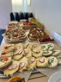 Auf einem Tisch stehen mehrere Tablets mit belegten Brötchen, Brezeln und Obst. Im Hintergrund mehrere Thermoskannen.