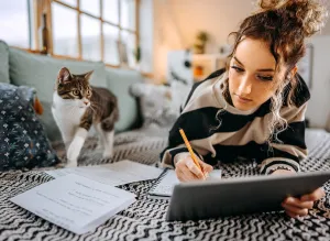 Eine junge Frau liegt mit ihrer Katze entspannt auf dem Sofa und liest im Tablet. Um Sie herum liegen Zettel mit Notizen.